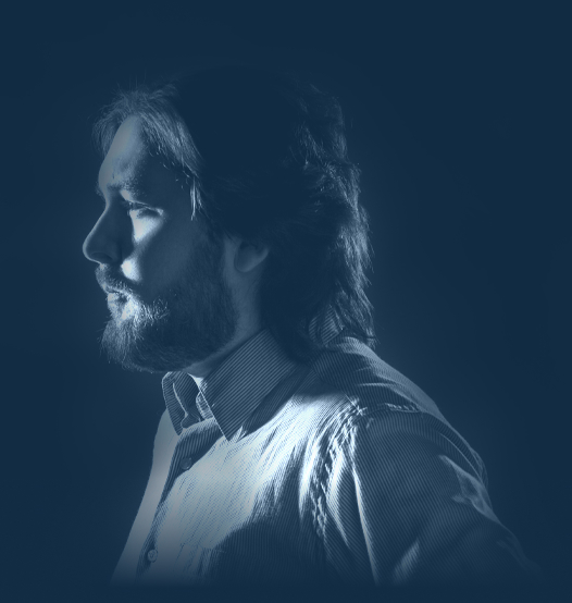 Man i profil med axellångt hår och skägg, i randig skjorta och blåtonat ljus.